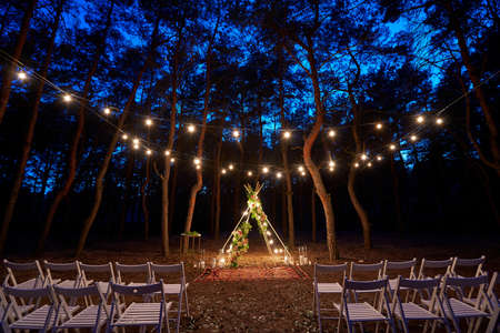 Festive string lights illumination on boho tipi arch decor on outdoor wedding ceremony venue in pine forest at night. Vintage string lights bulb garlands shining above chairs at summer rural wedding.の写真素材