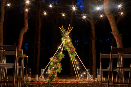 Festive string lights illumination on boho tipi arch decor on outdoor wedding ceremony venue in pine forest at night. Vintage string lights bulb garlands shining above chairs at summer rural wedding.の写真素材