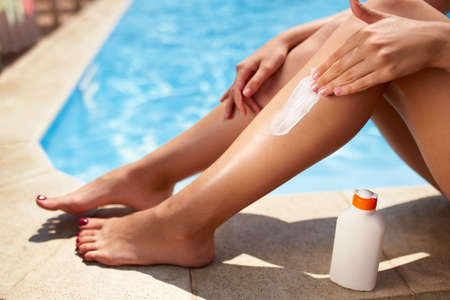 Slim woman applies suntan cream on legs sitting near swimming pool. Girl puts water resistant sunscreen lotion from a bottle on body on poolside deck. Skin burn and anti cancer care concept.の写真素材