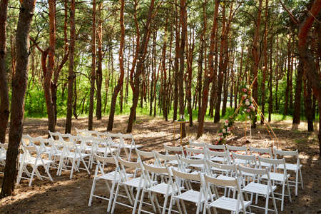 Beautiful bohemian tipi arch decoration on outdoor wedding ceremony venue in pine forest with cones. Chairs, floristic flower compositions of roses, carpet, string fairy lights. Summer rural wedding.の写真素材