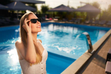 Pretty woman relaxing and sun bathing near swimming pool at luxury spa. Beautiful serene young female model in sunglasses on holiday travel resort enjoying vacation. Sun protection concept.の写真素材