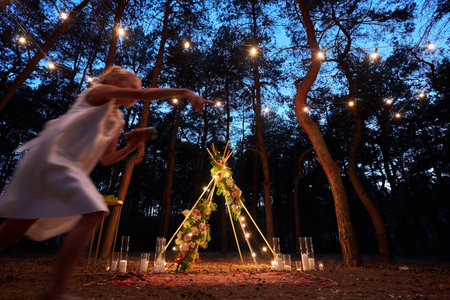 Festive string lights illumination on boho tipi arch decor on outdoor wedding ceremony venue in pine...