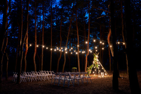 Festive string lights illumination on boho tipi arch decor on outdoor wedding ceremony venue in pine...