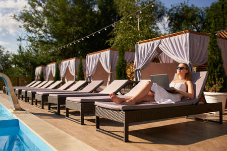 Self employed woman telecommuting with team on project from tropical island resort. Freelancer businesswoman doing remote work with laptop on swimming pool beach bed at spa. Girl tanning and relaxing.の写真素材