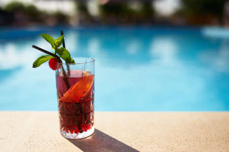 Glass of strawberry cocktail decorated with mint and cherry on swimming pool deck. Refreshing alcohol drink on water background with copy space. Vacation on resort and spa concept.の写真素材