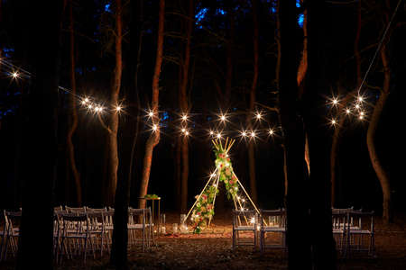Festive string lights illumination on boho tipi arch decor on outdoor wedding ceremony venue in pine...