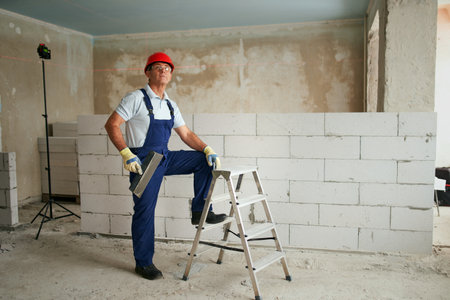 Professional construction worker in uniform standing with spatula on step ladder. Portrait of contractor in hardhat, overalls near masonry indoors. Handyman services for renovation with copy space.の写真素材