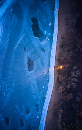Aerial top view from above of lonesome car at frozen sea coastline with headlights on. Lonely road trip traveller enjoys ocean ice floes scenics from vehicle on beach. Winter evening vivid blue colorsの写真素材