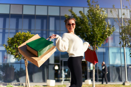 Happy woman with shopping bags walking in city from mall. Excited girl enjoys purchases on black friday sale. Female posing on camera with full paper bags. Consumerism concept.の写真素材