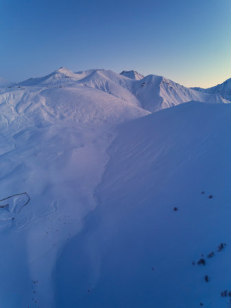 Aerial of snowy mountain slopes full of powder for freeride at ski resort on winter sunrise. Mountains range of backcountry covered with virgin snow. Caucasus peaks skyline with twilight afterglow.の写真素材