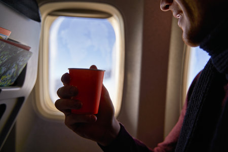 Smiling airline passenger with a paper cup of coffee looking ahead in airplane window. Man drinks tea onboard. Male holds beverage or water during flight.の写真素材