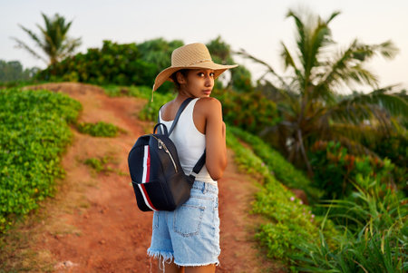 Multiethnic woman in straw hat enjoys tropical vacation travel hike. Black female with backpack sightseeing on scenic location. Pretty lady standing on island on sunrise. Girl in the sun for suntanの写真素材