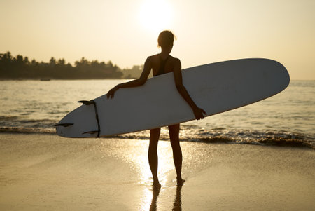 African american woman walking with surfboard on ocean beach. Black female surfer posing with surf board. Pretty multiethnic girl goes on surfing session, enjoys sun at tropical location at sunrise.の写真素材