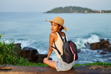 Multiethnic woman in straw hat enjoys tropical vacation sitting on bench at cliff with ocean view. Black female with backpack sightseeing on scenic location. Girl tanning on island on sea sunrise.の写真素材