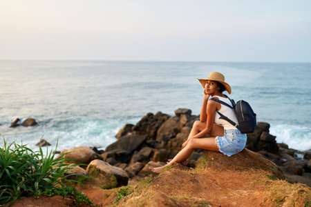 Multiethnic woman in straw hat enjoys tropical vacation sitting on rocky cliff with sea view. Black female with backpack sightseeing on scenic location. Pretty lady tanning on island on ocean sunrise.の写真素材