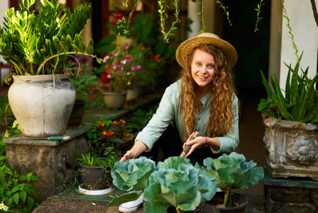 Young woman sitting in garden planting vegetables smiles and looks at camera. Junior female gardener with gardening tool in her orchard taking care of vegetables. Farming and gardening conceptの写真素材