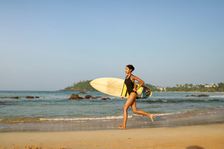 African american woman running with surfboard on ocean beach. Black female surfer with surf board. Pretty multiethnic girl goes on surfing session on tropical location at sunny sunrise.の写真素材