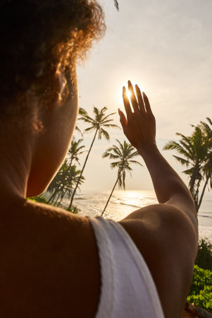 Silhouette of delicate feminine hand covering the sun. Black woman covers sun with hand and fingers closeup shot from back. African young woman stretches arm and fingers to sun and plays with lightの写真素材