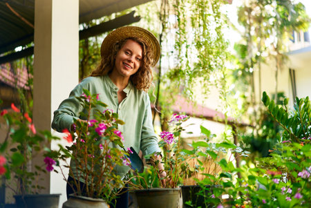 Young woman standing in garden planting flowers in pots smiles and looks at camera. Junior female gardener with gardening tool in orchard taking care of potted plants. Farming and gardening conceptの写真素材