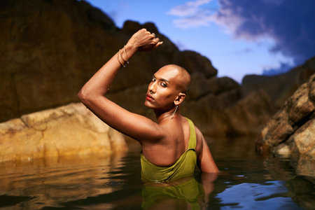 Non-binary black person stands chest deep in the middle of still water inside scenic creek at night. Lgbtq ethnic graceful fashion model poses immersed in crystal clear pond at exotic location at duskの写真素材