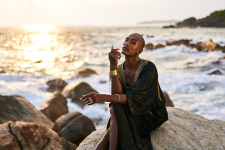 Non-binary ethnic fashion model in dress, brass jewelry sits on rocks by ocean. Trans sexual black person with rings, nose-ring, bracelets, earrings in posh clothes poses in tropical seaside location.の写真素材