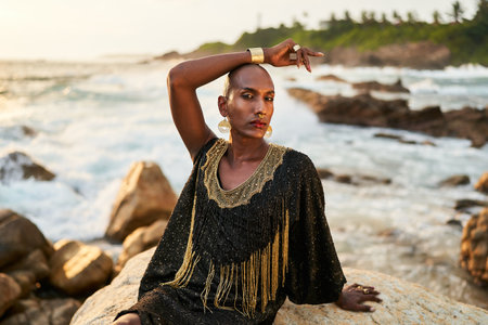 Androgynous ethnic fashion model in luxury dress, jewelry sits on rocks by ocean. Gay black person in jewellery, posh clothes poses gracefully in tropical seaside location. Pride LGBTQ bipoc concept.の写真素材
