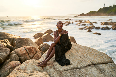 Epatage lgbtq black man poses on scenic ocean beach looks at camera demonstrates jewellery . Androgynous ethnic fashion model in posh dress looks at camera, sits on stone inの写真素材