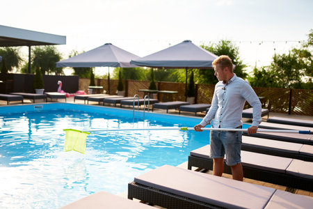 Man with skimmer net equipment cleaning swimming pool from rubbish. Service and maintenance of the...