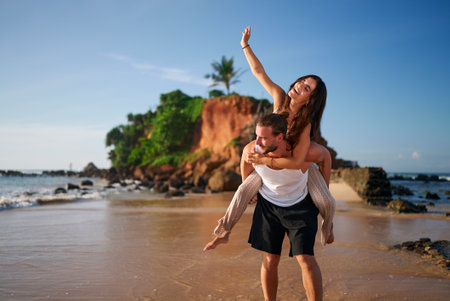 Joyful couple enjoys a romantic piggyback ride on a serene beach. Man carries girlfriend, playful adventure by sea. Relaxed travel, loving pair explores tropical shore, happy woman raises hand.の写真素材