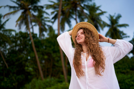 Joyful young woman enjoys a tropical getaway among lush palm trees, holding onto her straw hat,...