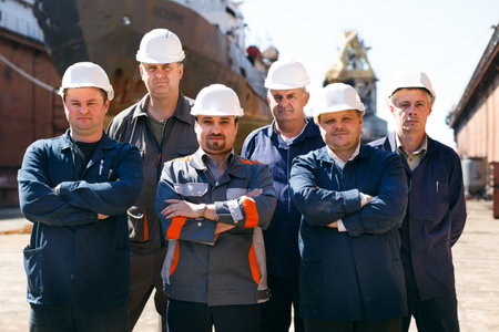 Shipyard workers at float dock wearing hard hats, stand arms-crossed. Male repair team ready for maintenance work on vessel. Maritime industry, professional ship fix group, outdoor labor day scene.の写真素材