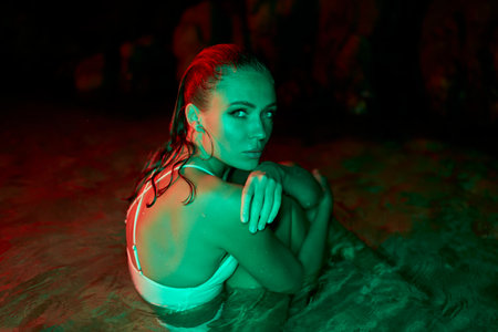 Woman immerses in lagoon, nocturnal setting with red and green illumination. Reflective mood, contemplative female submerged, water surrounds soothingly in dark. Pensive state, watery embrace at nightの写真素材