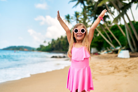 Happy little girl in pink dress, sunglasses smiles, arms raised in joy on sandy tropical beach, with clear blue sky, coconut palms, playful atmosphere in sunny daylight for family vacation memories.の写真素材