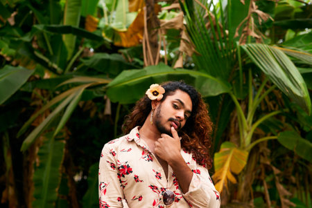 Elegant gay man with flower in hair poses thoughtfully in garden. Exotic male in floral shirt embraces self-love, LGBTQ pride against green foliage. Inclusive portrait fashion, diversity themes.の写真素材