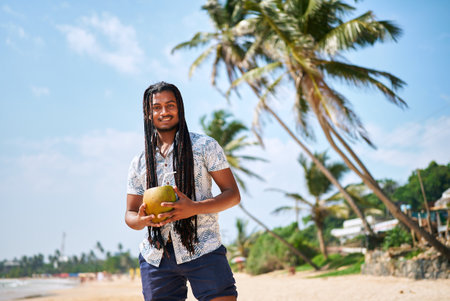 Bipoc man with dreads enjoys fresh coconut on sunny beach. Relaxed traveler sips natural drink, tropical vibe. Palm trees, blue sky frame exotic escape. Solo journey, wellness refreshment by sea.の写真素材