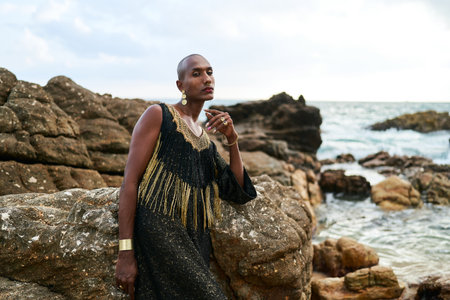 Queer black person in luxury dress sits on rocks in ocean. Lgbtq ethnic fashion model wearing jewellery dressed in posh gown poses gracefully in tropical seaside location portrait. Pride monthの写真素材