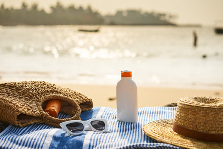 Sun protection gear laid out on seaside day. Straw hat, sunglasses, sunscreen bottle on blue towel....