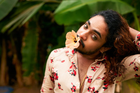 South Asian man with expressive eyes, flower in mouth, models in tropical setting. Flamboyant gay individual explores femininity, cultural identity through bold fashion, natural backdrop.の写真素材