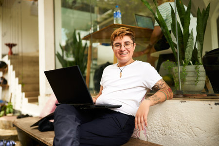 Transgender Gen Z freelancer smiles while working on laptop at eco-friendly coworking space. Inked, digital nomad in casual clothes, emphasizing diversity, inclusivity in modern remote work settings.の写真素材