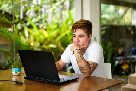 Transgender pro works on laptop in home office setting. Confident and focused individual busy with work task, surrounded by green plants, embodying workplace inclusion and gender identity acceptance.の写真素材