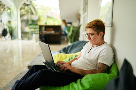 Transgender professional works on laptop, seated on green bean bag, in casual office space. Relaxed environment, light-filled room, modern work life balance, inclusion in workplace highlighted.の写真素材