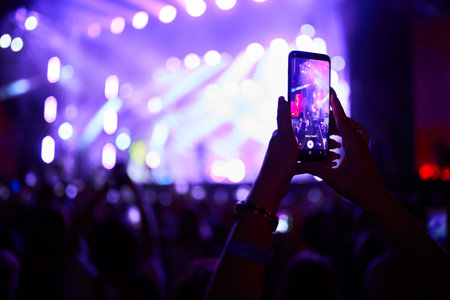 Excited audience at music fest, vibrant lights illuminate stage, fan records live performance on smartphone, friends enjoy night out, band plays in background, festival vibe in summer evening.の写真素材
