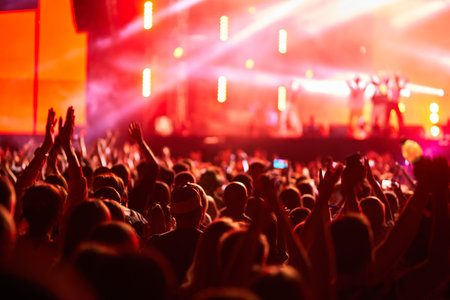 Energetic audience at outdoor live concert at sunset. Fans raise hands, enjoy music at summer festival. Stage lights illuminate excited crowd. Event in full swing with vibrant energy, community vibe.の写真素材