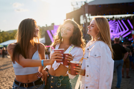 Group of women toast with cups at beachside music event. Smiling friends enjoy summer festival vibe. Party atmosphere with dancing, laughter by sea. Casual attire, sunset light, joyful celebration.の写真素材
