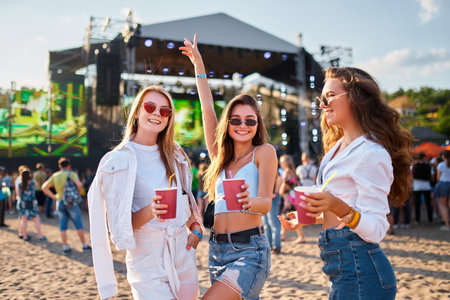 Girls toast with cold drinks at beach music fest in summer, smiling, wearing sunglasses, with stage, people in background. Celebration, fun, youth lifestyle. Females enjoying party, ocean nearby.の写真素材