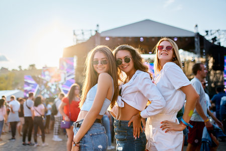 Group of three young women enjoy summer music festival on beach. Friends dance, laugh in sun, near sea stage. Party atmosphere with crowd in background. Casual wear, sunglasses, golden hour fun.の写真素材