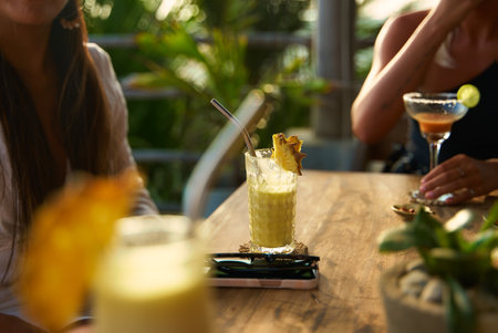 Two women enjoy drinks at outdoor cafe. Pineapple cocktail and margarita on wooden table. Relax in tropical setting with lush plants. Social dining and summer vibes.の写真素材
