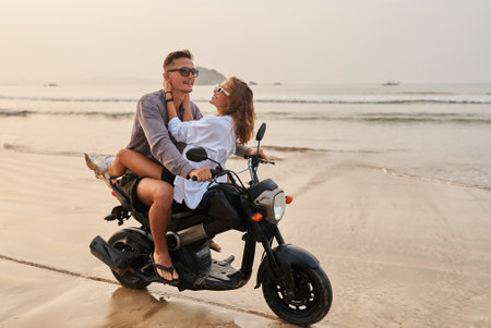 Couple enjoys romantic ride on motorbike along beach. Smiling lovers on motorcycle by ocean at sunset. Travel, love, adventure with scooter on sandy shore. Man, woman explore new places.の写真素材