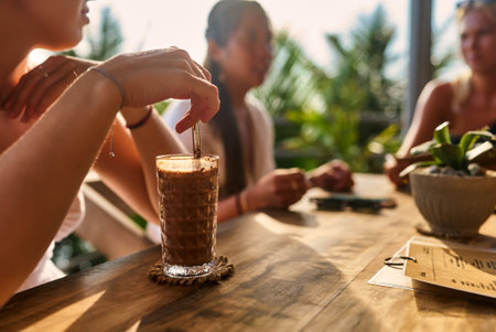 Group of friends relax in outdoor cafe with iced coffee drinks. Sunlit patio with rich plants. People chat, enjoy beverages. Casual meeting or social gathering.の写真素材