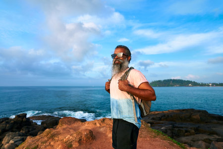 Old man hikes coastal cliff with backpack under blue sky. Senior enjoys active lifestyle. Adventurous male explores retirement travel. Embraces golden years. Happy elder near ocean.の写真素材